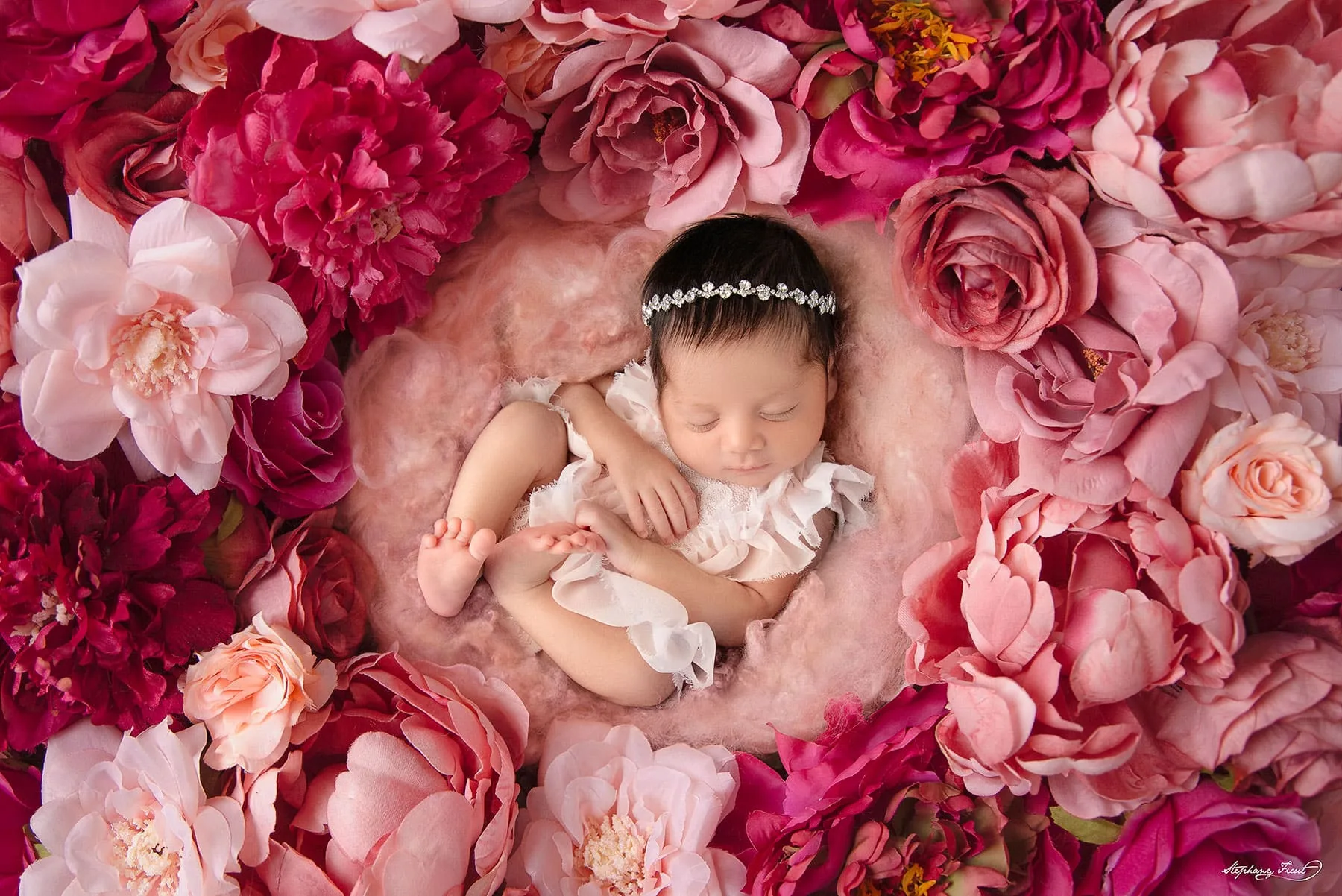 A newborn baby is peacefully sleeping on a fluffy pink blanket, surrounded by vibrant, colorful flowers. The backdrop is adorned with lush, blooming petals, enhancing the soft, delicate atmosphere of the scene. The baby is dressed in a simple, light pink outfit, blending harmoniously with the floral theme. The overall image exudes a warm and cheerful ambiance, capturing the innocence and beauty of the newborn. This setup by Stephany Ficut showcases a creative and charming newborn photoshoot theme.